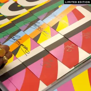 A hand rests on a vibrant grid of Poster Lakwena, 2025, Limited Edition prints—each signed, numbered, and displaying bold geometric patterns in red, yellow, green, blue, and black, with a LIMITED EDITION label in the top right corner.