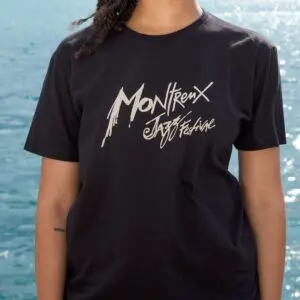 Une personne se tient au bord de l'eau et porte le T-Shirt Montreux Jazz Festival, Noir, avec un lettrage blanc frappant "Montreux Jazz Festival" sur un tissu noir. Seule la partie supérieure du corps est visible.