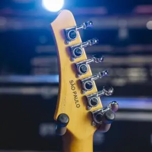 Gros plan de l'arrière d'une tête de guitare Montreux Guitar Collection São Paulo, Série limitée, avec des chevilles d'accord, étiquetée SÃO PAULO, sur un fond flou et des lumières de scène brillantes.