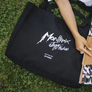 Un sac Shopping Bag, Montreux Jazz, portant le nom du festival en blanc sur noir, repose sur de l'herbe verte à côté d'une personne tenant un éventail pliant.