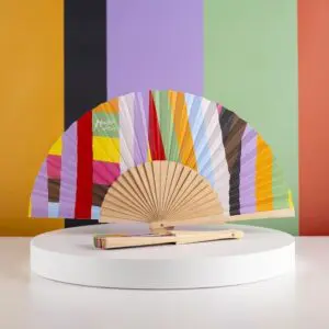MJF25-Shooting-Lakwena-eventail-2.jpg L'Éventail Lakwena, un éventail à main avec des rayures multicolores et un manche en bois, est présenté ouvert sur un support blanc à côté de l'éventail fermé assorti, sur fond de rayures orange, noires, vertes et pêche.