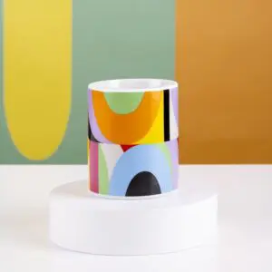 The Mug Lakwena, a vibrant cylindrical mug with bold abstract geometric patterns, rests on a white circular pedestal. Behind it, large curved shapes in yellow, green, orange, and teal evoke the lively Montreux Jazz spirit.