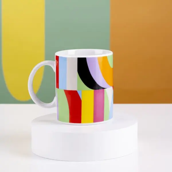 The Mug Lakwena, a white mug with a vibrant abstract geometric pattern, rests on a round white pedestal. Bold yellow and white shapes on green and orange walls evoke energetic Montreux Jazz festival vibes.