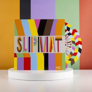 A colorful vinyl record and cover stand on a white pedestal. Both feature bold, geometric stripes in various bright colors, with the word “SLIPMAT” in multicolored letters on the cover against a striped background.