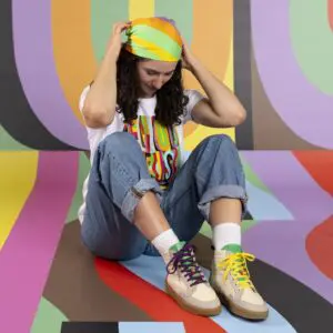Une personne est assise sur un sol et un fond colorés et rayés. Elle porte un jean retroussé, un foulard arc-en-ciel, un tee-shirt blanc avec un texte multicolore et des baskets crème aux lacets dépareillés.