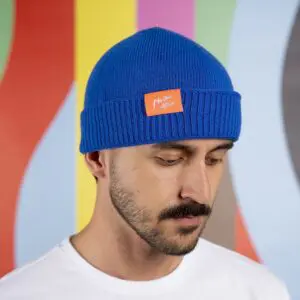 Un homme portant un bonnet bleu du Montreux Jazz Festival avec un écusson orange regarde vers le bas, arborant une moustache, une courte barbe et une chemise blanche. Le fond rayé vibrant et ondulé fait écho à l'esprit vif de Montreux.