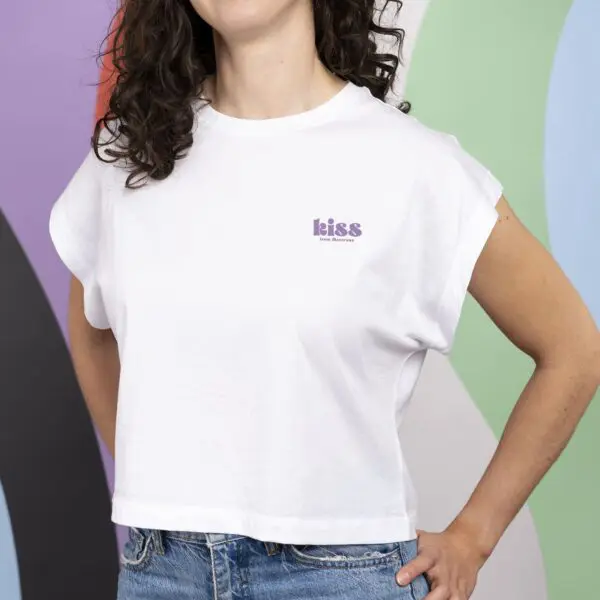 Une personne aux cheveux bruns ondulés porte le T-shirt Crop, Kiss - un crop top blanc avec des manches roulées, "kiss" en texte violet sur la poitrine gauche, et un jean bleu - sur un fond coloré, abstrait, inspiré de Montreux.