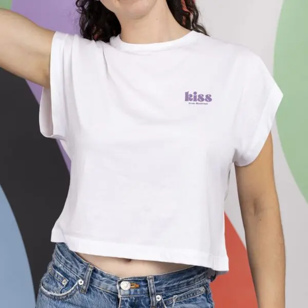 A person wears the white T-shirt Crop, Kiss with rolled sleeves and "kiss" printed in purple on the chest, paired with blue jeans, standing before a vibrant, abstract background.