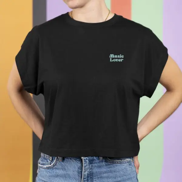 A person wearing the T-shirt Crop, Music Lover—a black tee with small white "Music Lover" text—paired with blue jeans, stands before a colorful striped background. Their head is partially out of frame.