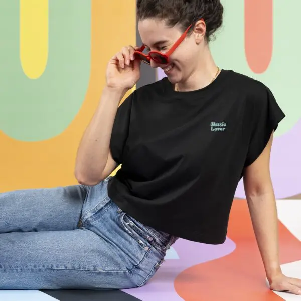 A woman with dark hair in a bun, wearing red sunglasses and blue jeans, smiles on a colorful abstract background. She is dressed casually in the "T-shirt Crop, Music Lover.