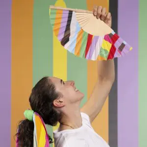 A smiling person with a rainbow-striped scarf in their hair holds up the Hand Fan Lakwena at the Montreux Festival, with vertical pastel stripes in the background. They look up at the colorful fan, inspired by Lakwena.