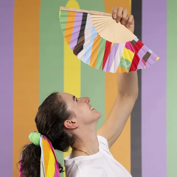 A smiling person with a rainbow-striped scarf in their hair holds up the Hand Fan Lakwena at the Montreux Festival, with vertical pastel stripes in the background. They look up at the colorful fan, inspired by Lakwena.