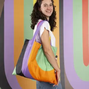 A smiling woman with curly hair holds the Bag LOQI Lakwena, a vibrant tote with orange, green, purple, and black curves, standing in front of an abstract background inspired by Montreux Jazz festival artistry.