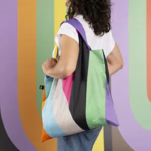 Une personne aux cheveux noirs et bouclés, vêtue d'une chemise blanche et d'un jean, porte le sac fourre-tout Sac LOQI Lakwena. L'arrière-plan présente des rayures verticales multicolores qui font écho au motif du sac, inspiré du jazz de Montreux.