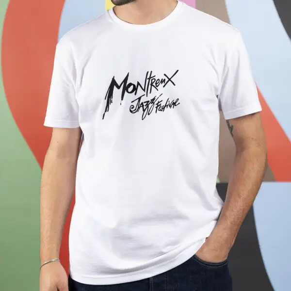 Une personne portant le T-Shirt Montreux Jazz Festival, Blanc avec "Montreux Jazz Festival" dans une police artistique se tient sur un fond géométrique coloré.
