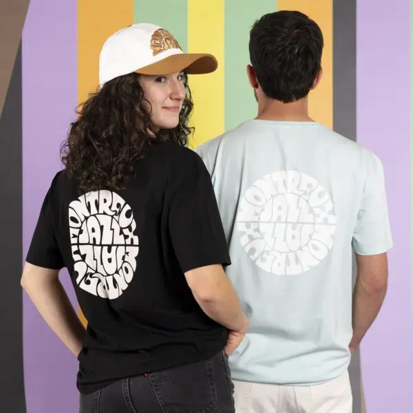 Deux personnes se tiennent dos à dos, face à la caméra. L'une porte un T-Shirt Vintage Montreux Jazz Festival, Noir, et une casquette blanche ; l'autre porte une chemise bleu clair. Leurs tee-shirts comportent un texte rond et gras, avec un mur rayé coloré derrière eux.