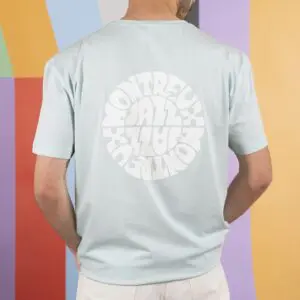 Une personne vêtue d'un T-Shirt Vintage Montreux Jazz Festival, Bleu ciel, avec un logo circulaire blanc du festival au dos, se tient devant un fond géométrique vibrant.