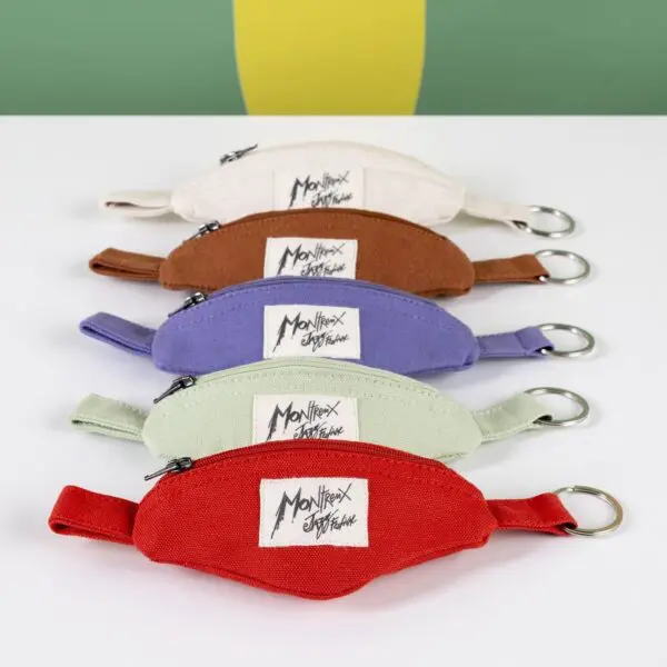 Cinq Porte-clés Mini Sac Banane colorés avec fermetures éclair et porte-clés sont alignés. Chaque pochette est étiquetée Montriex Est. France et disponibles en beige, marron, violet, vert et rouge - parfait pour les fans du Montreux Jazz.