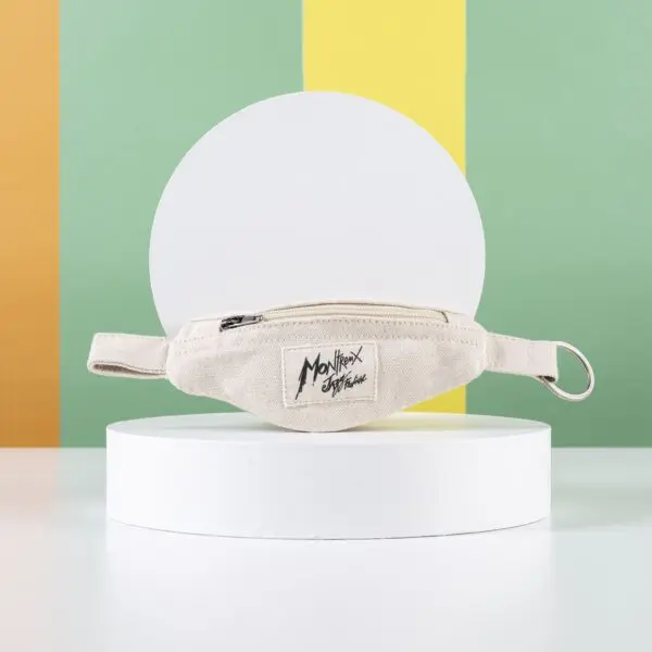 Le Porte-clés Mini Sac Banane, un sac de taille en tissu beige avec une fermeture éclair noire et un écusson Mon Nrex Originals, est présenté sur un socle rond et blanc avec un porte-clés banane, sur un fond rayé vert, jaune et orange.