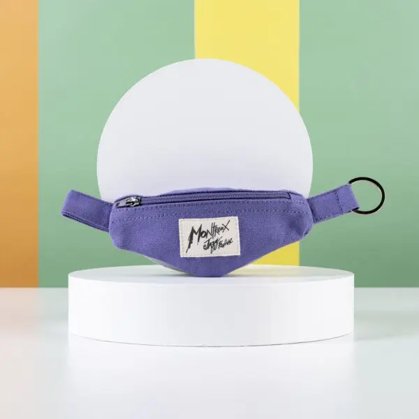 Une petite pochette violette à fermeture éclair portant l'étiquette Monthex Signature est posée sur un support circulaire blanc, accompagnée d'un Porte-clés Mini Sac Banane, sur un fond de rayures verticales colorées jaunes, orange et vertes.
