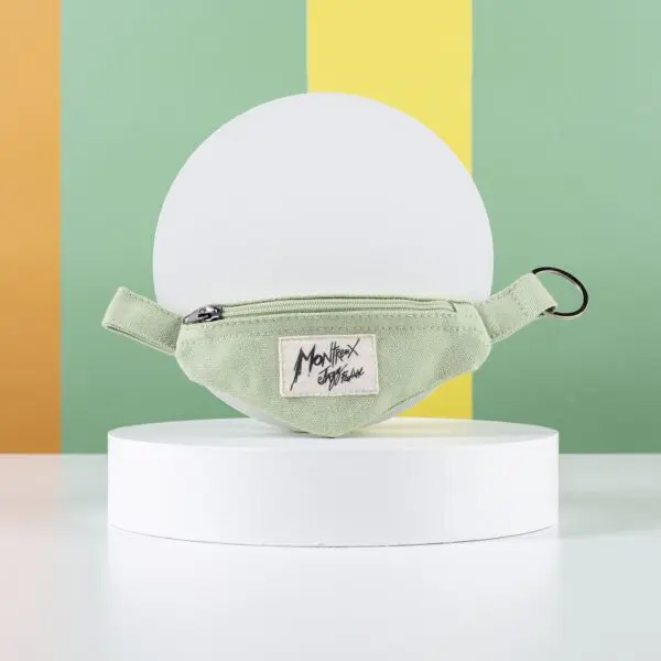 Le Porte-clés Mini Sac Banane, une petite pochette zippée vert clair avec un porte-clés, est posé sur un support rond blanc. Elle affiche un écusson "Montrex Style" sur un fond rayé coloré - idéal pour les amateurs de jazz de Montreux.