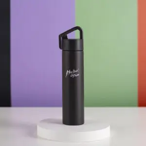 Une Mini Bouteille Isotherme 180ml noire avec un couvercle à poignée et le texte "Monthex Lifestyle" se trouve sur une plateforme ronde blanche, sur un fond de bandes verticales violettes, noires, vertes et rouges.