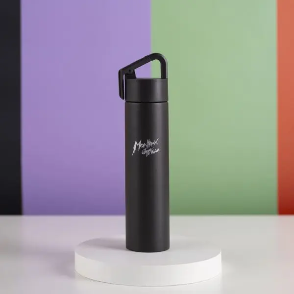 Une Mini Bouteille Isotherme 180ml noire avec un couvercle à poignée et le texte "Monthex Lifestyle" se trouve sur une plateforme ronde blanche, sur un fond de bandes verticales violettes, noires, vertes et rouges.