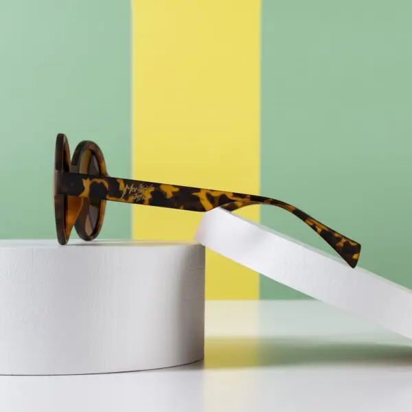 Side view of Fly Amsterdam Light Tortoiseshell Glasses resting on white cylindrical platforms, set against a green and yellow striped background.