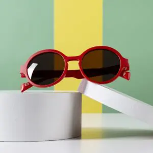 Les lunettes de soleil Lunettes Fly Amsterdam Pêche, rondes et rouges avec des verres foncés, sont présentées sur des plateformes cylindriques blanches devant un fond rayé vert et jaune vibrant inspiré du Montreux Jazz.