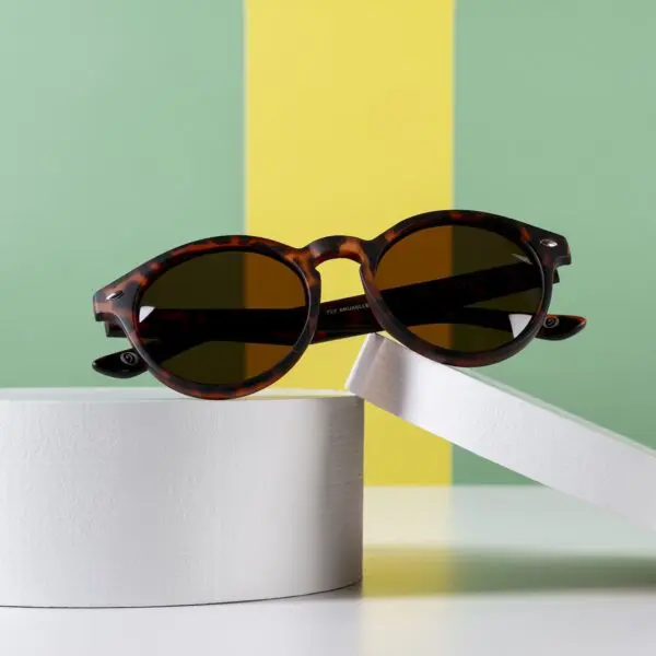Une paire de lunettes de soleil Lunettes Fly Bruxelles Écaille foncée avec des verres foncés est présentée sur une plate-forme circulaire blanche sur un fond rayé vert et jaune, reflétant l'esprit vibrant du Montreux Jazz.