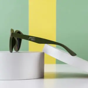 Une paire de lunettes de soleil Lunettes Fly Bruxelles Kaki repose sur deux plateformes cylindriques blanches sur fond vert et jaune. La branche des lunettes de soleil est ornée d'un logo blanc.