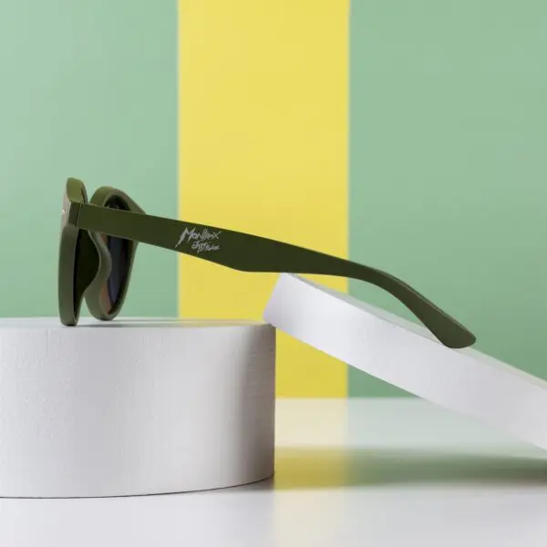 Une paire de lunettes de soleil Lunettes Fly Bruxelles Kaki repose sur deux plateformes cylindriques blanches sur fond vert et jaune. La branche des lunettes de soleil est ornée d'un logo blanc.