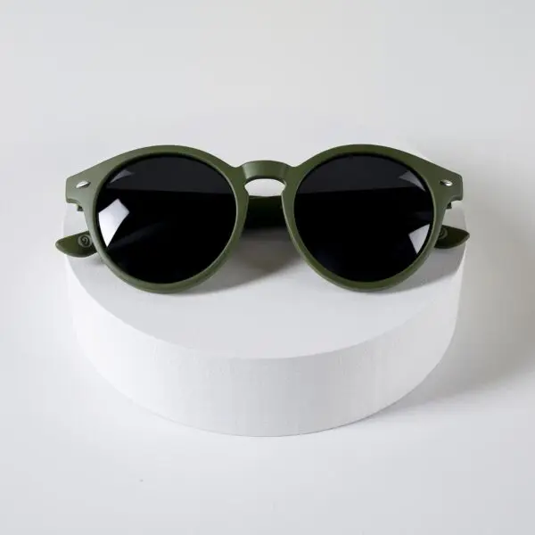 Des lunettes de soleil rondes aux verres foncés et à la monture vert olive - Lunettes Fly Bruxelles Kaki- sont présentées sur un socle cylindrique blanc sur un fond blanc uni.