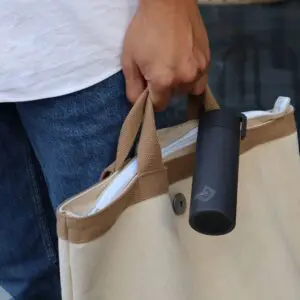 Une personne en jean bleu et chemise blanche porte un sac fourre-tout beige avec des poignées marron. Une Mini Bouteille Isotherme 180ml noire et élégante est accrochée à l'une des poignées.
